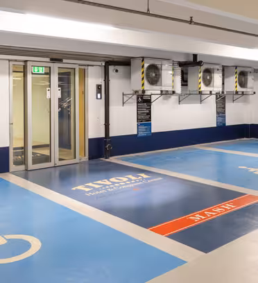 Tilgængelige parkeringspladser i en garage med blå kørestolssymboler. På et skilt på jorden står der "Tivoli". Der er vægskilte med information og dør til venstre. Overhead er der ventilationsaggregater.