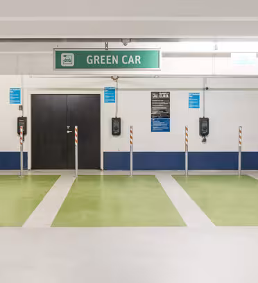 Parkeringsplads til elbiler med grønmalede pladser under et "GRØN BIL" skilt. Der er monteret ladestandere på væggen mellem parkeringsbanerne og flere små skilte med instruktioner.