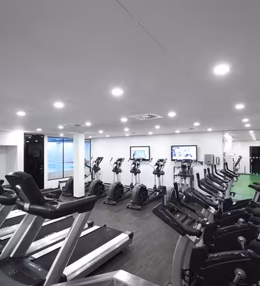 Et moderne fitnesscenter med flere løbebånd og stationære cykler opstillet på et mørkt trægulv. To tv-skærme er monteret på bagvæggen. Rummet er godt oplyst med lyse loftslys, og en del af væggen er beklædt med spejle.