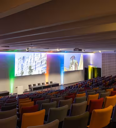 Et tomt auditorium med farverige sæder ud mod en scene. Scenen har et podium og seks stole. Væggene viser store skærme, der viser arkitektoniske billeder. Belysningen kaster forskellige farver hen over væggene.