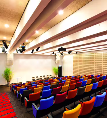 Et moderne auditorium med farverige, polstrede sæder arrangeret i rækker. Loftet har forede paneler med forsænket belysning. En lille scene med en talerstol og et par planter er synlig foran. Væggene er dekoreret med akustikfliser.