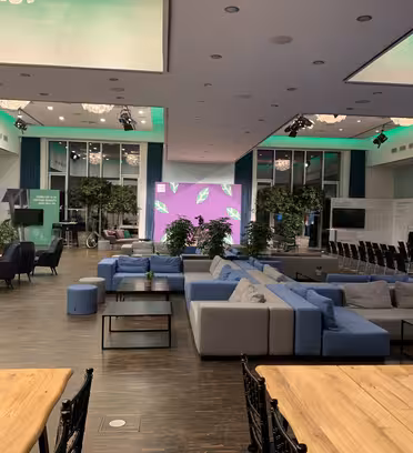Et moderne mødelokale med trægulve og et stort opholdsområde med blå og grå sofaer. Stole er arrangeret i teaterstil i baggrunden, og grønt lys fremhæver loftet. Store digitale skærme viser abstrakte mønstre.