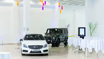 En hvid Mercedes-Benz A-klasse og en sort Mercedes-Benz SUV vises i et lyst showroom med hvide vægge og gulve. Fra loftet hænger farverige pendler, og runde borde med hvide duge er spredt rundt.
