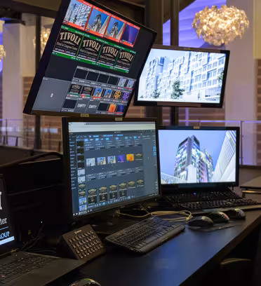 Et kontrolbord med flere skærme, der viser billeder og videoer. Opsætningen inkluderer en skærm, der viser "TIVOLI Hotel & Congress Center" og forskellige billeder. Moderne belysningsarmaturer hænger ovenover i et rummeligt rum.