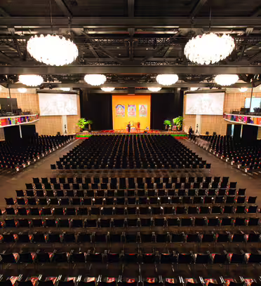 Rummeligt auditorium med rækker af tomme sorte stole ud mod en scene med farverig vægkunst og to store skærme. Elegante lysekroner hænger fra loftet. Balkon siddepladser er synlige ovenfor.