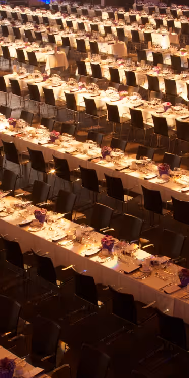 En ekspansiv festsal fyldt med rækker af lange, formelle spiseborde sat med hvide duge, tallerkener, glasvarer og lilla blomsterdekorationer. Rækker af mørke stole står langs hvert bord. Rummet er elegant oplyst.