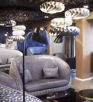 Moderne lounge med stilfulde, buede siddepladser i grå nuancer. Unikke pendellamper hænger fra loftet. Indretningen har en blanding af teksturer og mønstre, hvilket skaber en smart og hyggelig atmosfære. En drikkevarestation er synlig i baggrunden.
